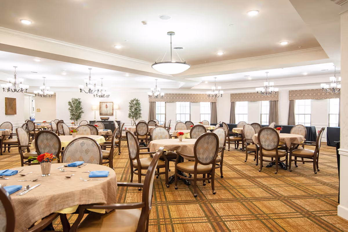 A spacious dining room with multiple round tables covered with beige tablecloths, each set with blue napkins, silverware, and small flower arrangements. The room features patterned carpet flooring, upholstered chairs with wooden frames, large windows with curtains, and elegant chandeliers hanging from the ceiling. There are decorative plants and framed artwork on the walls.