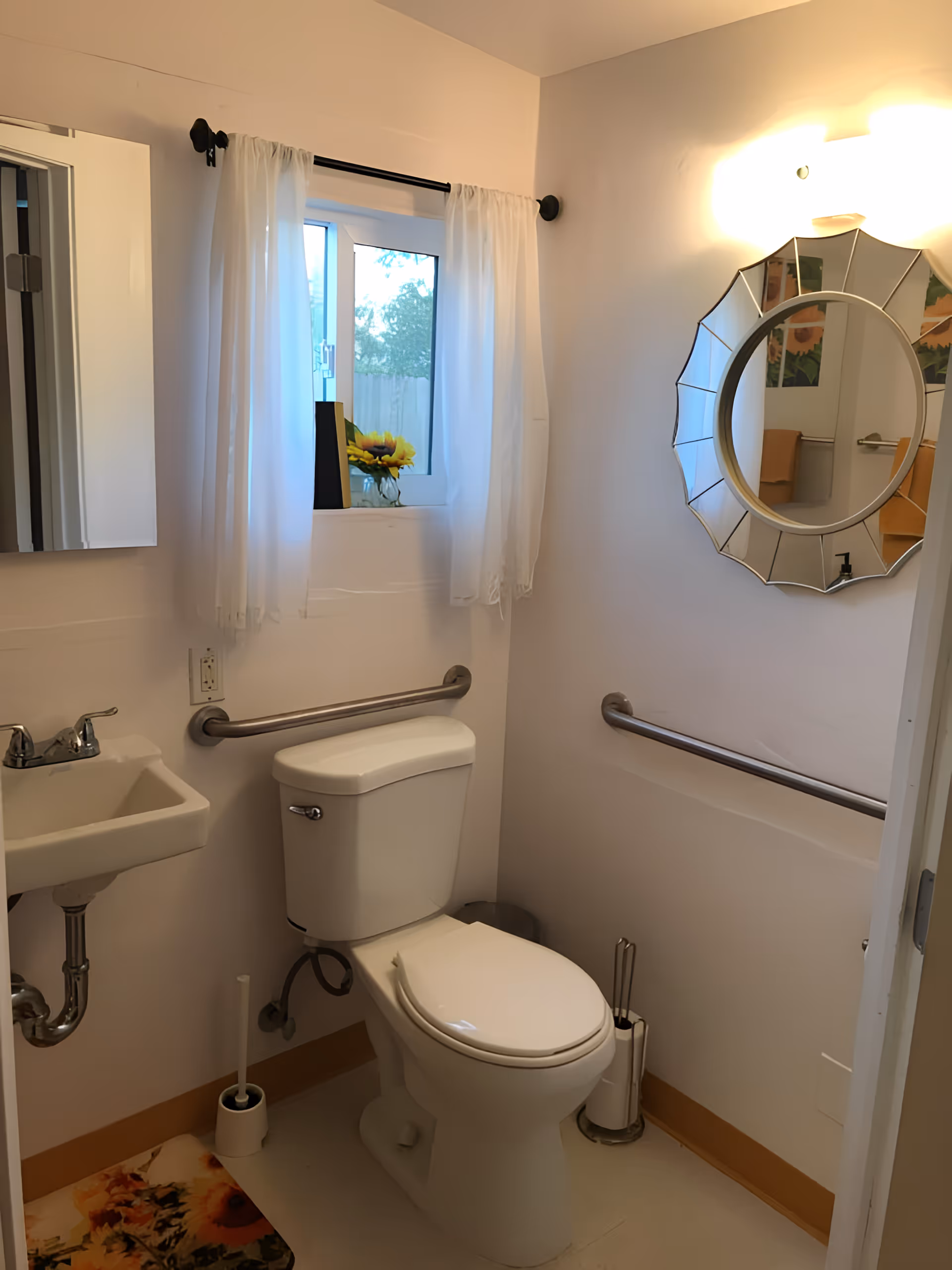 Small accessible bathroom with a toilet, wall-mounted sink, grab bars, a decorative round mirror, and a curtained window with sunflowers on the sill.