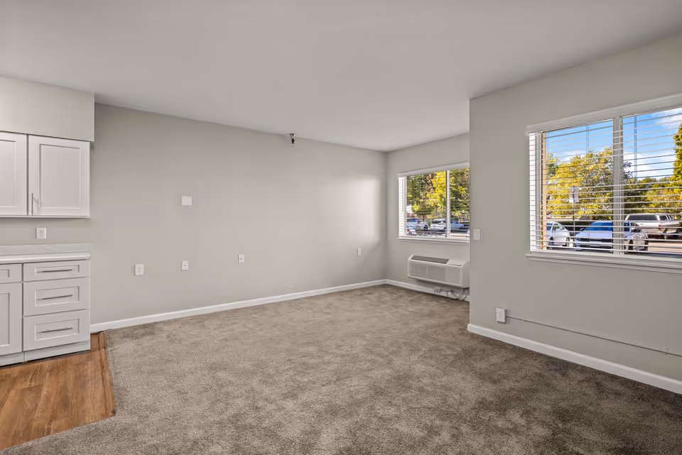 Empty room with beige carpet and light gray walls, featuring two windows with white blinds showing a parking lot and trees outside. On the left side, there are white kitchen cabinets and a small section of wood flooring.