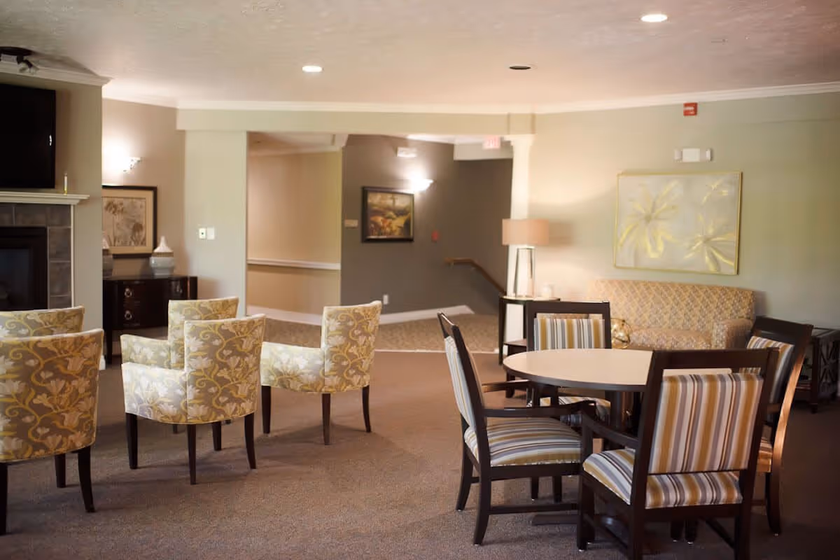 A cozy senior living common area with a round table surrounded by four striped cushioned chairs in the foreground. Behind the table are four floral patterned armchairs arranged in front of a fireplace with a mounted TV above it. The room has soft lighting, neutral-colored walls, a beige carpet, and decorative artwork on the walls.
