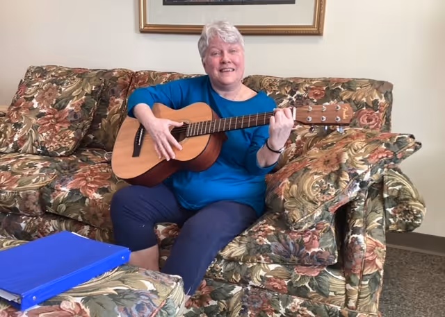 An elderly woman with short white hair sitting on a floral-patterned couch playing an acoustic guitar. She is wearing a blue long-sleeve shirt and dark pants. There is a matching floral-patterned ottoman with a blue binder on it in front of her. The room has beige walls and a framed picture hanging above the couch.
