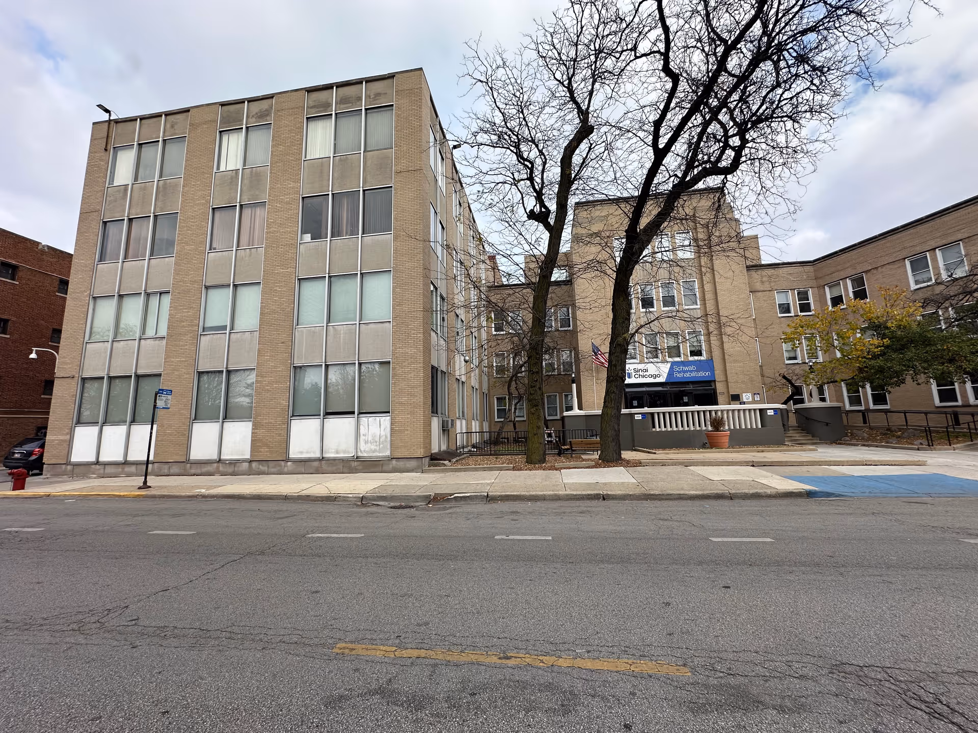 Exterior view of Schwab Rehabilitation Center SNU, a multi-story beige brick building with numerous windows, a sidewalk, and leafless trees in front under a cloudy sky.