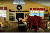 A cozy living room decorated for the holidays with a large poinsettia plant in the foreground, a fireplace with a wreath above it, shelves with holiday decorations, and comfortable seating including armchairs. The room has large windows with red valances letting in natural light.