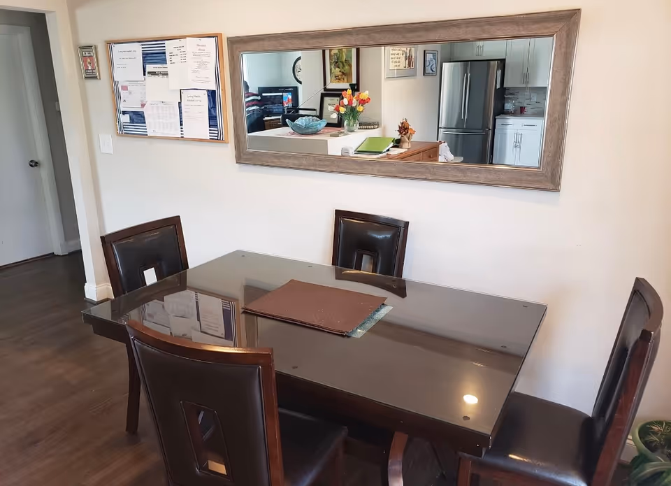 Dining area with a glass-top table and four chairs beneath a large wall mirror reflecting the kitchen.