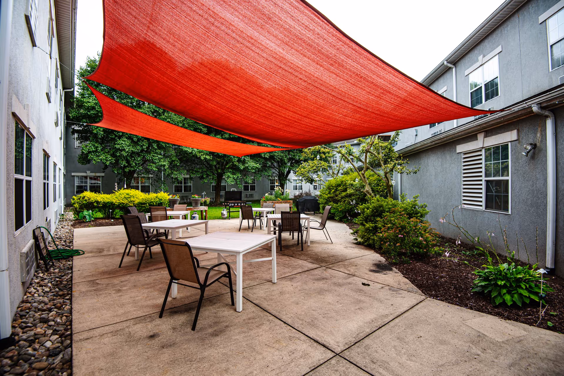 Outdoor patio area at Orwigsburg Nursing and Rehabilitation Center with several tables and chairs arranged on a concrete surface. Red fabric sun shades are stretched overhead, providing shade. The patio is surrounded by greenery including bushes, trees, and landscaped garden beds, with the facility building visible on both sides.