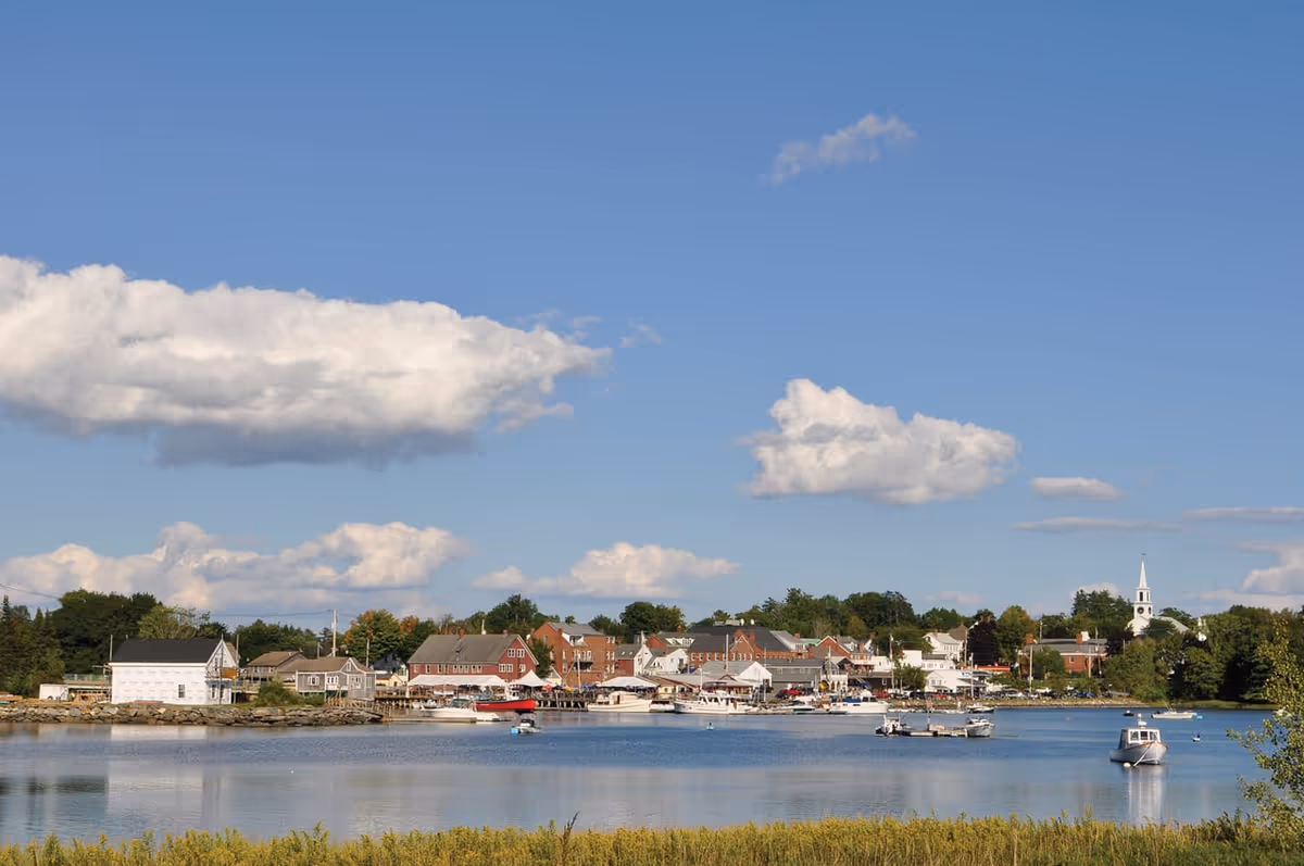 Small coastal town skyline with waterfront houses and boats on calm water under a blue sky with clouds.