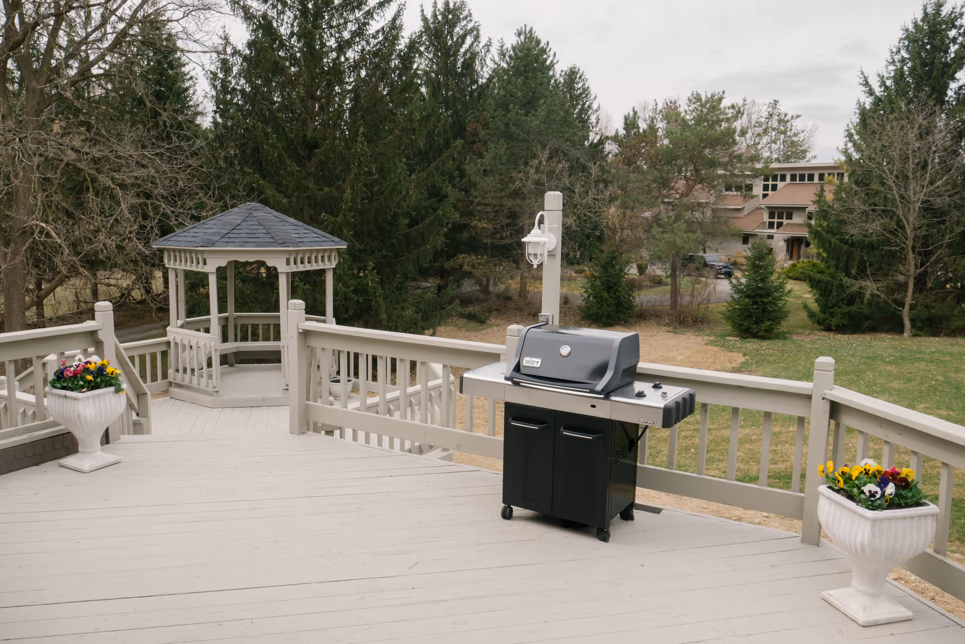Outdoor wooden deck area with a black Weber Spirit gas grill, two white planters with colorful flowers, a small gazebo, and trees in the background.