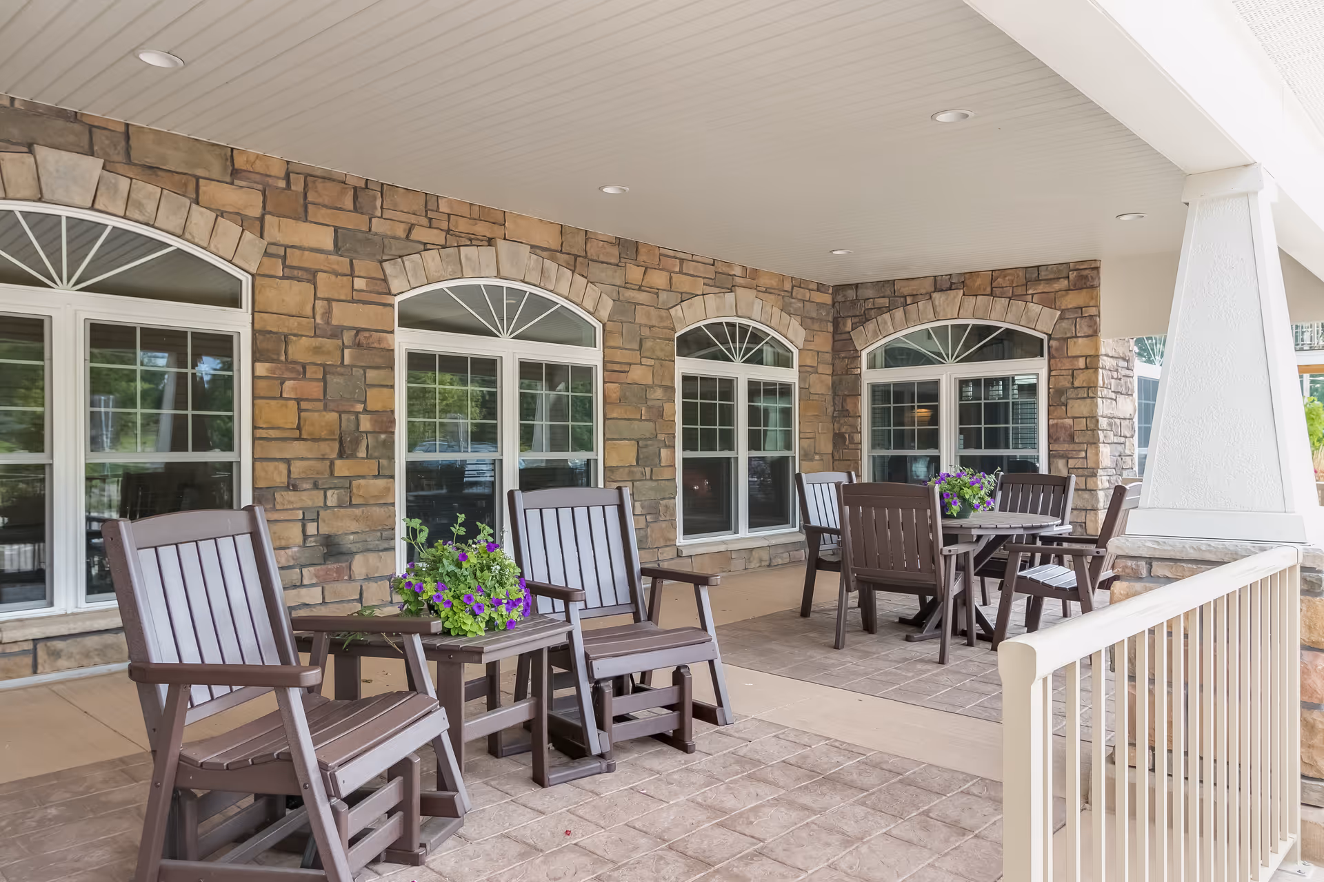 Covered outdoor patio area with stone walls and multiple windows. The patio is furnished with brown wooden chairs and tables, some with potted purple flowers on them. The ceiling is white with recessed lighting, and there is a white railing along the edge of the patio.