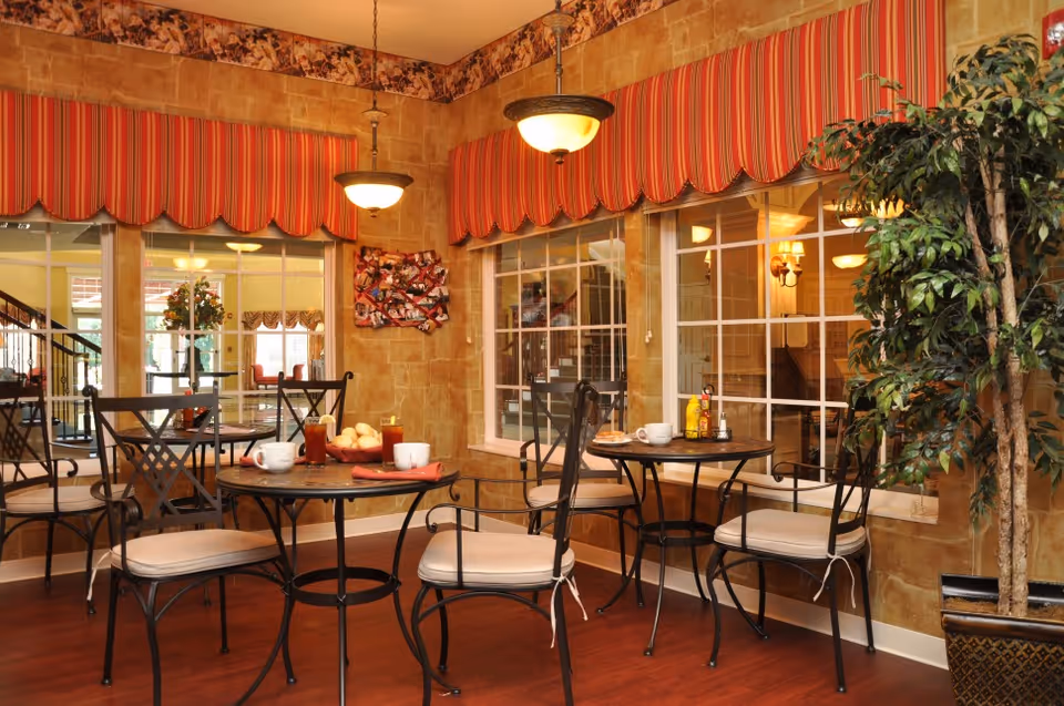 A cozy dining area with two round tables and black metal chairs with beige cushions. The tables have cups, glasses of iced tea, plates with food, and condiments. The walls are decorated with a warm stone-like texture and red striped valances above large windows. A potted plant is placed in the corner, and two hanging light fixtures illuminate the space.