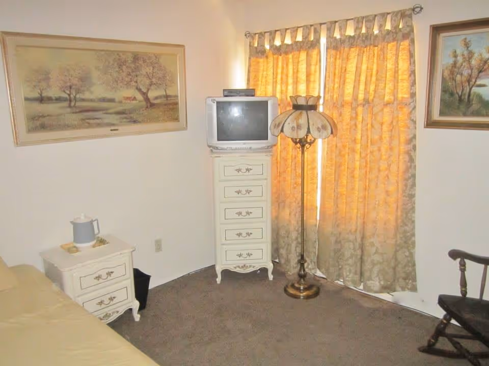A small living area with a beige couch on the left, a white nightstand with a pitcher and glass, a tall white chest of drawers with an old-style CRT television on top, a vintage floor lamp with a floral lampshade, and a window covered by yellow patterned curtains. There are two framed landscape paintings on the walls and a wooden rocking chair on the right.