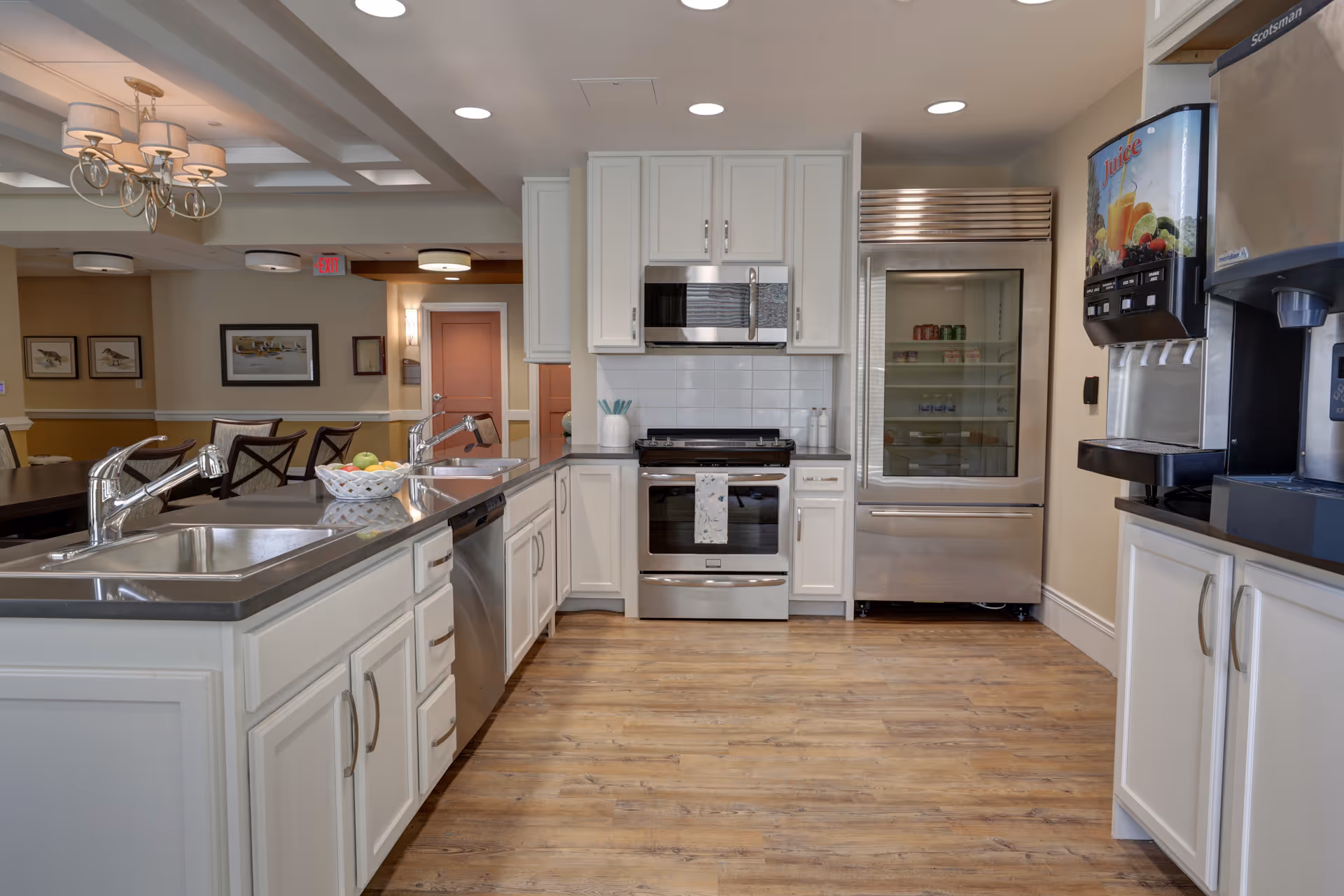 A bright communal kitchen with white cabinets, stainless-steel appliances, a central island with dual sinks, and a beverage dispenser, opening onto a dining area.