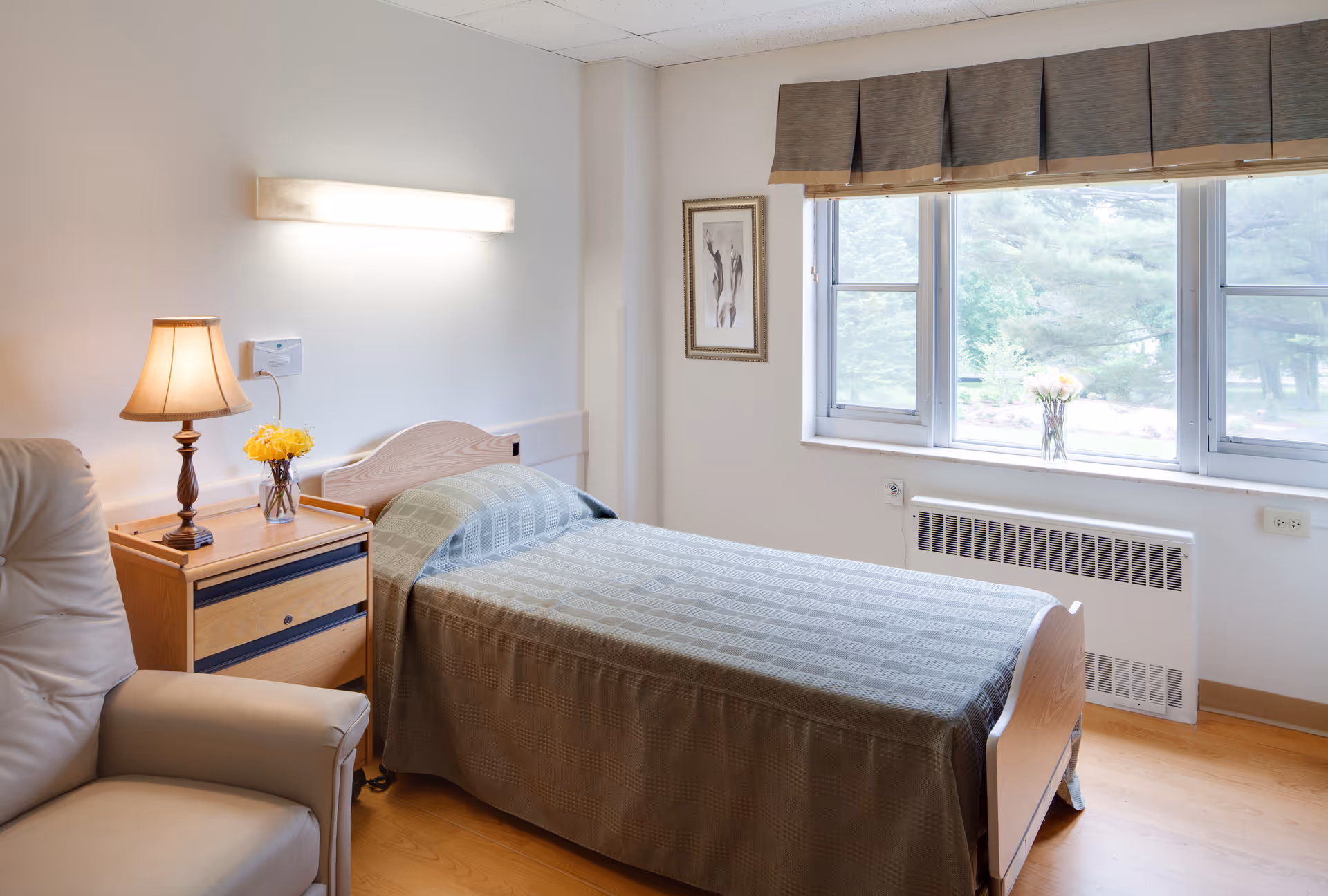 A simple, clean bedroom in a senior living facility with a single bed covered in a greenish-gray patterned bedspread. Next to the bed is a wooden nightstand with a lamp and a vase of yellow flowers. A comfortable beige armchair is partially visible in the foreground. The room has a large window with a brown valance, letting in natural light and showing greenery outside. A framed artwork hangs on the wall beside the window.