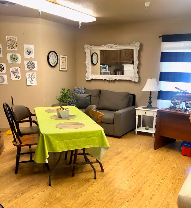 A cozy room with a green tablecloth-covered table surrounded by various chairs, a gray couch with pillows, a white side table with a lamp, a large ornate white-framed mirror on the wall, and a window with blue and white striped curtains. The wall also features a clock and several framed artworks.