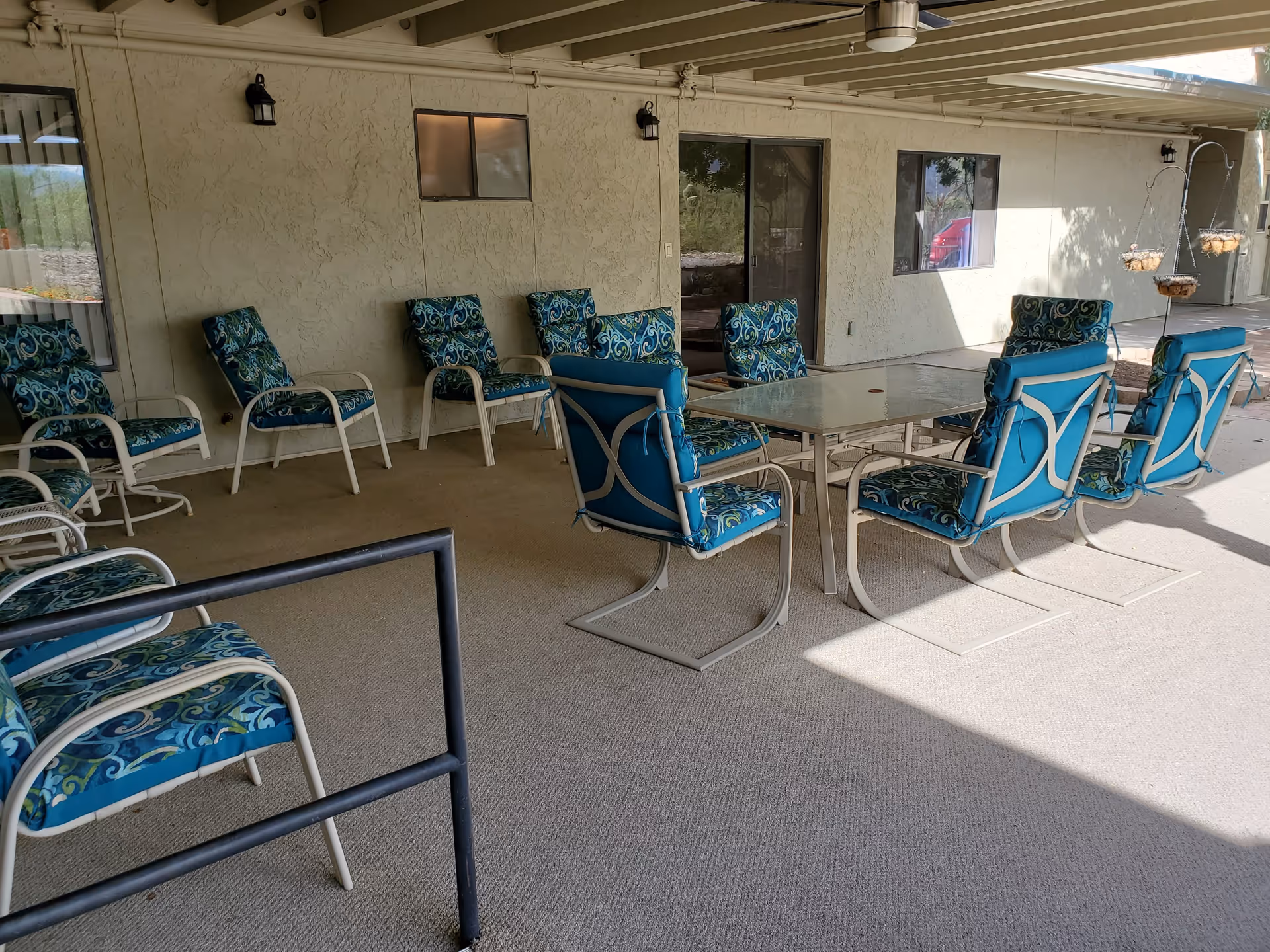 Covered outdoor patio area with multiple cushioned chairs arranged around a glass-top table and along the wall. The chairs have blue and green patterned cushions. The patio is attached to a building with beige stucco walls, windows, and a sliding glass door. There are hanging planters on the right side and outdoor light fixtures mounted on the wall.