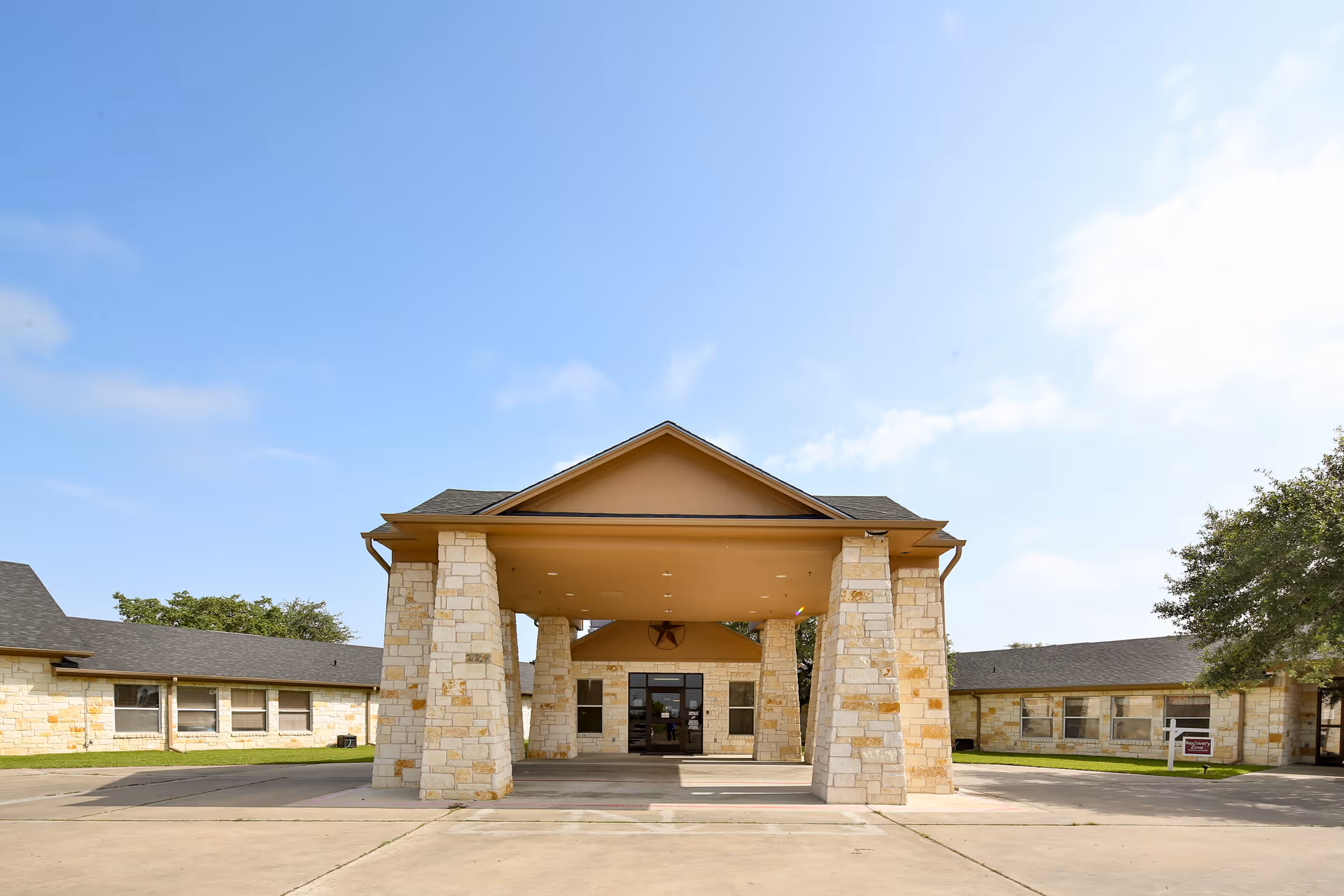 Front exterior of a single-story nursing facility with a covered stone-column entrance and flanking wings under a clear blue sky.