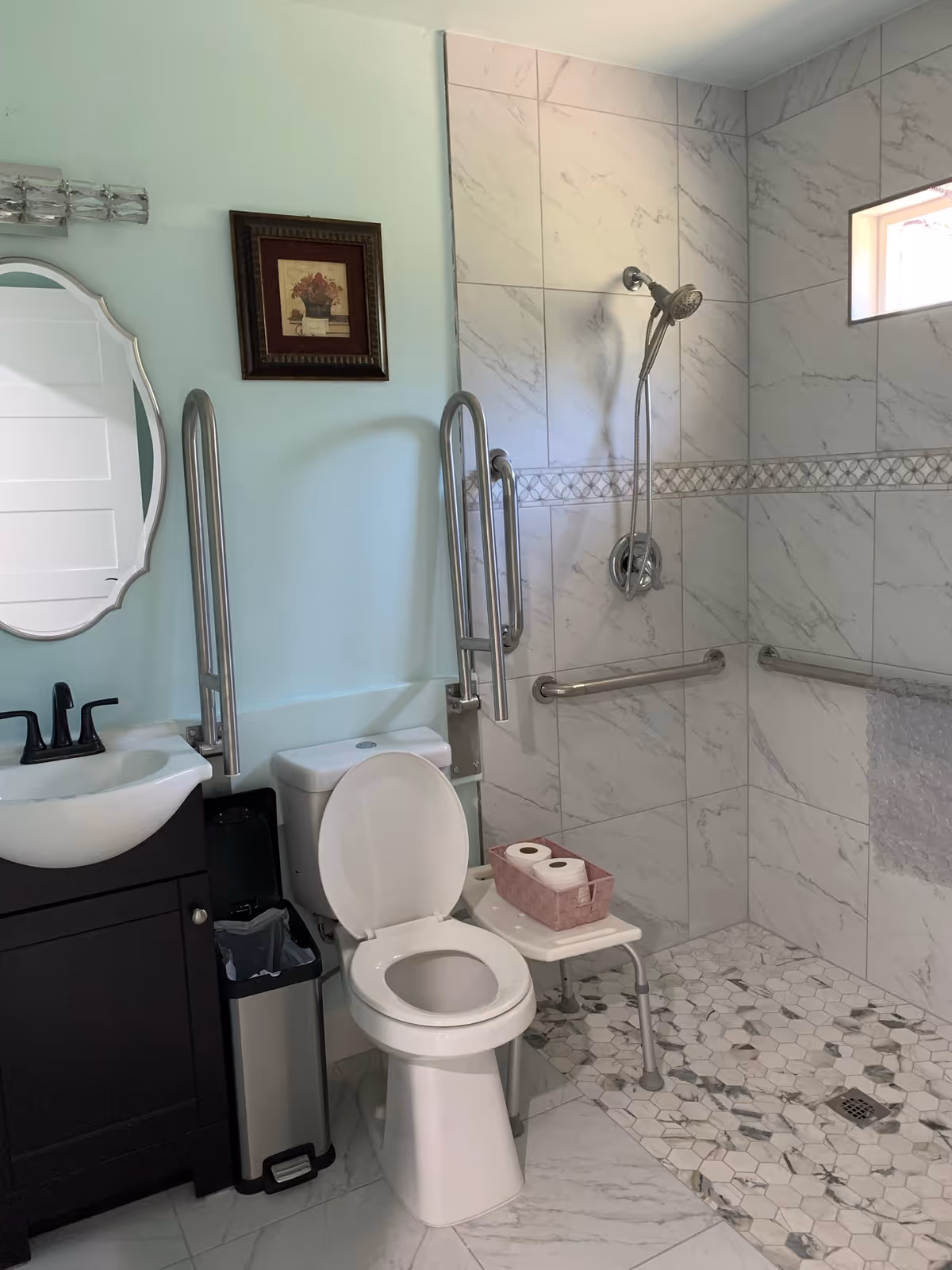 A bathroom designed for accessibility with a toilet equipped with grab bars on both sides, a small white bench holding a basket with toilet paper rolls, a walk-in shower with a handheld showerhead and grab bars, a dark brown vanity with a white sink and black faucet, a trash can, a decorative framed picture on the light blue wall, and a small window letting in natural light.