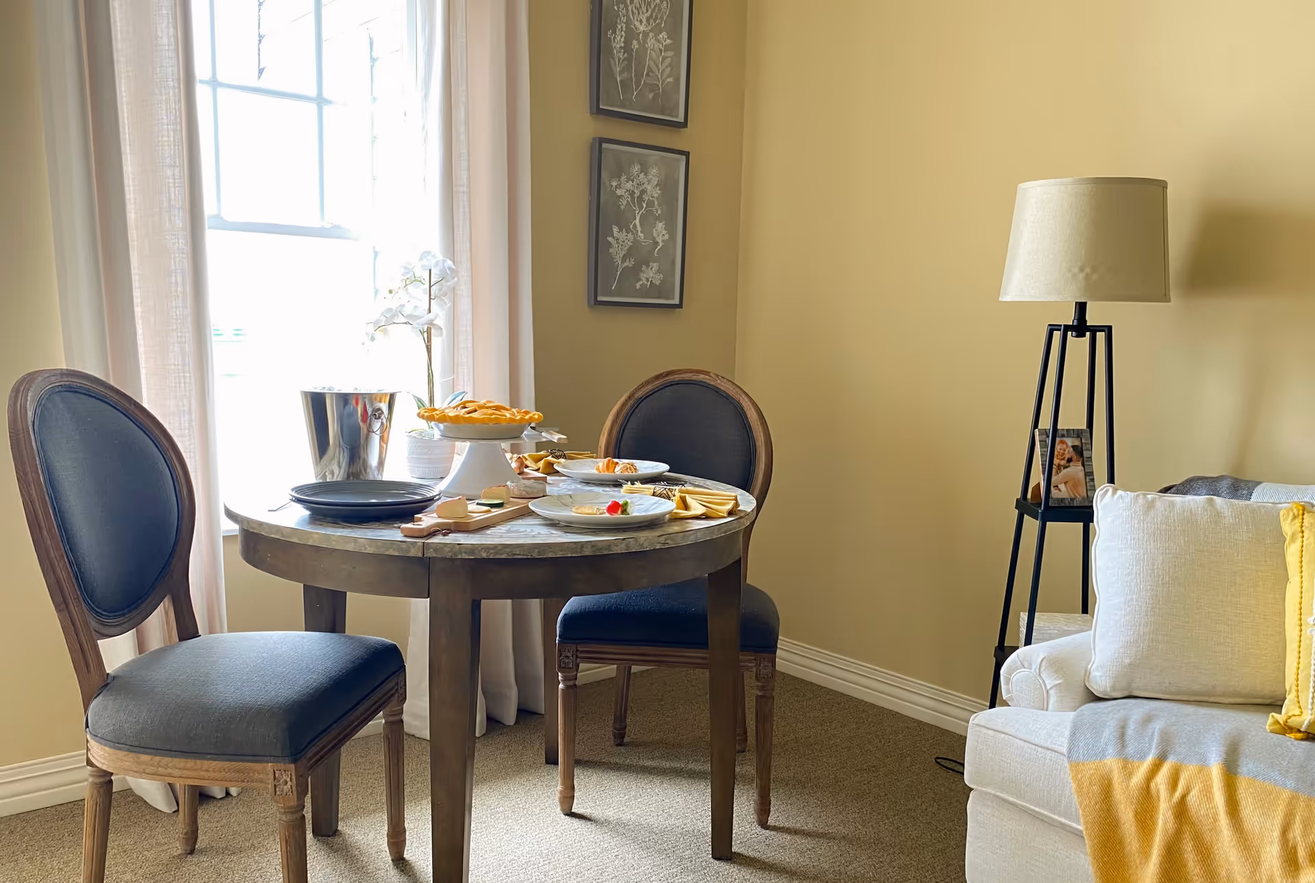A cozy dining area with a round wooden table set for a meal, featuring plates, a cheese board, and a pie on a cake stand. Two upholstered chairs with wooden frames are positioned around the table. A window with sheer white curtains allows natural light to brighten the room. To the right, there is a beige sofa with white and yellow pillows and a gray and yellow throw blanket. A black floor lamp with a beige shade and a small framed photo on its base stands next to the sofa. The walls are painted a soft yellow, and two framed botanical prints hang on the wall behind the table.