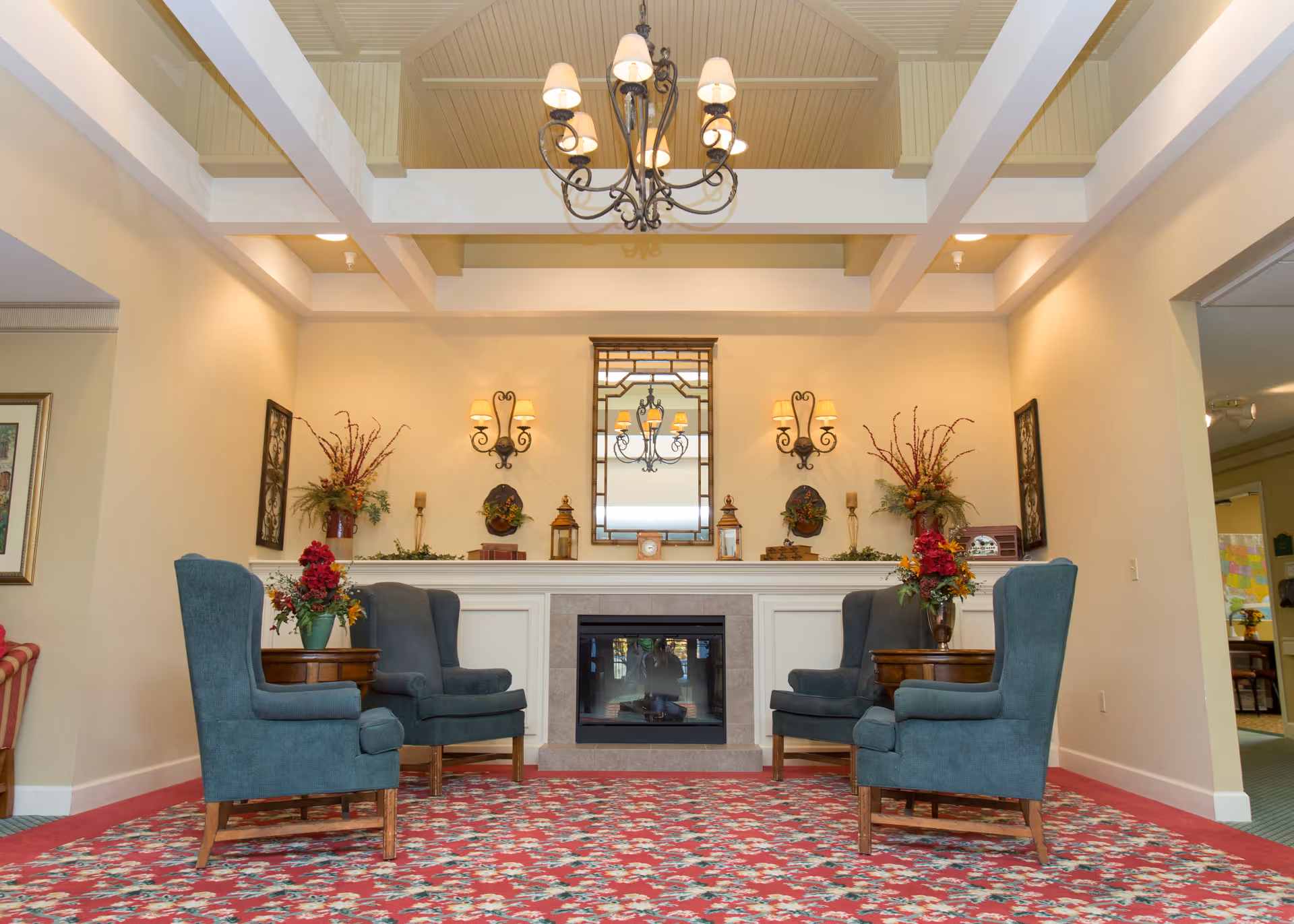 A cozy sitting area with four teal upholstered armchairs arranged around two wooden side tables with flower arrangements. The room features a fireplace with a decorative mantel, a large mirror above it, wall sconces with lampshades, and floral decorations. The ceiling has exposed beams and a chandelier with multiple lights. The floor is covered with a red floral carpet.