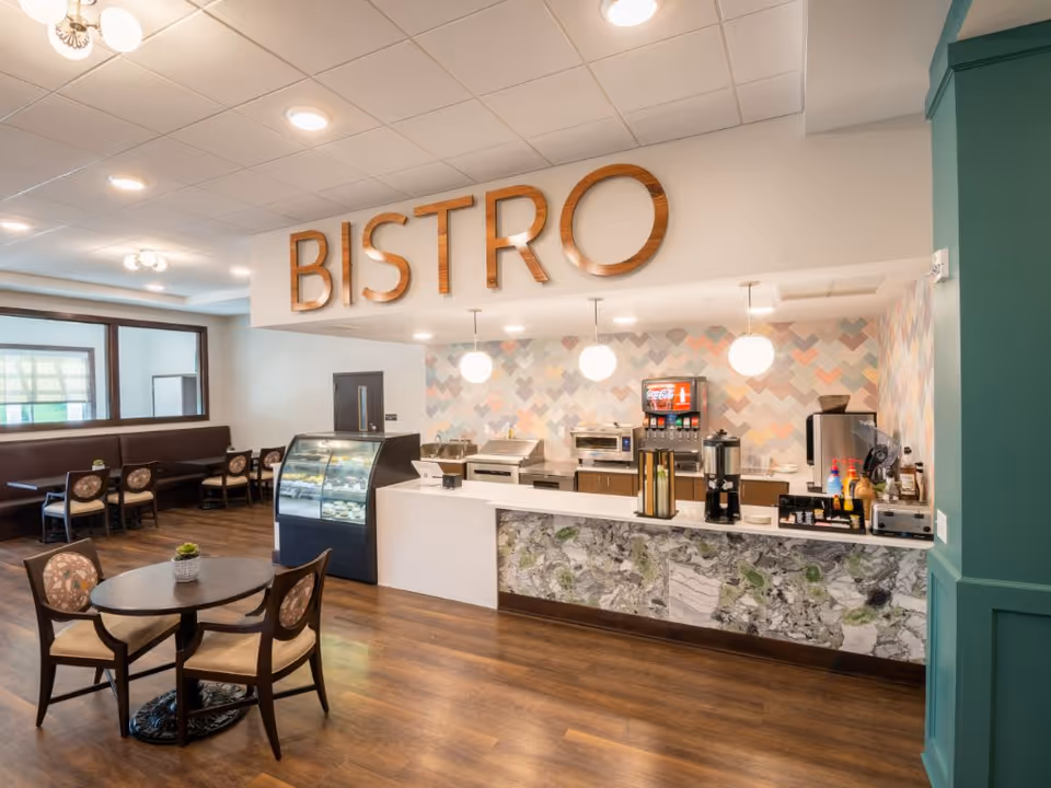 Interior view of a senior living facility bistro area with a serving counter, beverage dispensers, and a display case. There are round tables with chairs and booth seating along the wall. The word 'BISTRO' is prominently displayed in large wooden letters above the counter.