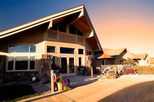 Exterior view of a senior living facility building with large windows, stone and wood siding, a sloped roof, and a patio area with outdoor seating and an umbrella. The scene is lit by warm sunlight under a clear sky.