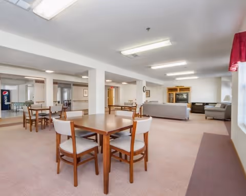 Spacious senior living common room with wooden dining tables and chairs and a seating area with couches and a television.