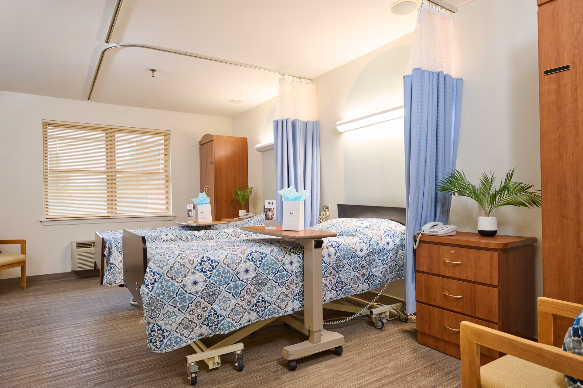 Bright shared care room with two hospital beds, blue patterned bedding, privacy curtains, wooden nightstands and a window.