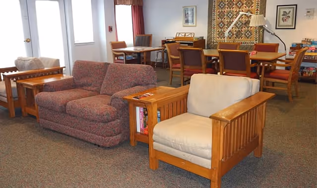 A cozy common area in an assisted living facility featuring a patterned fabric loveseat, a cushioned wooden armchair, wooden side tables with books, and several wooden tables and chairs in the background. The room has carpeted flooring, a quilt hanging on the wall, framed artwork, and a lamp.