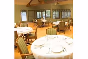A dining room with round tables covered with white tablecloths, set with plates, glasses, and silverware. The room has wooden floors, green cushioned chairs, large windows with valance curtains, and a high ceiling with exposed wooden beams.