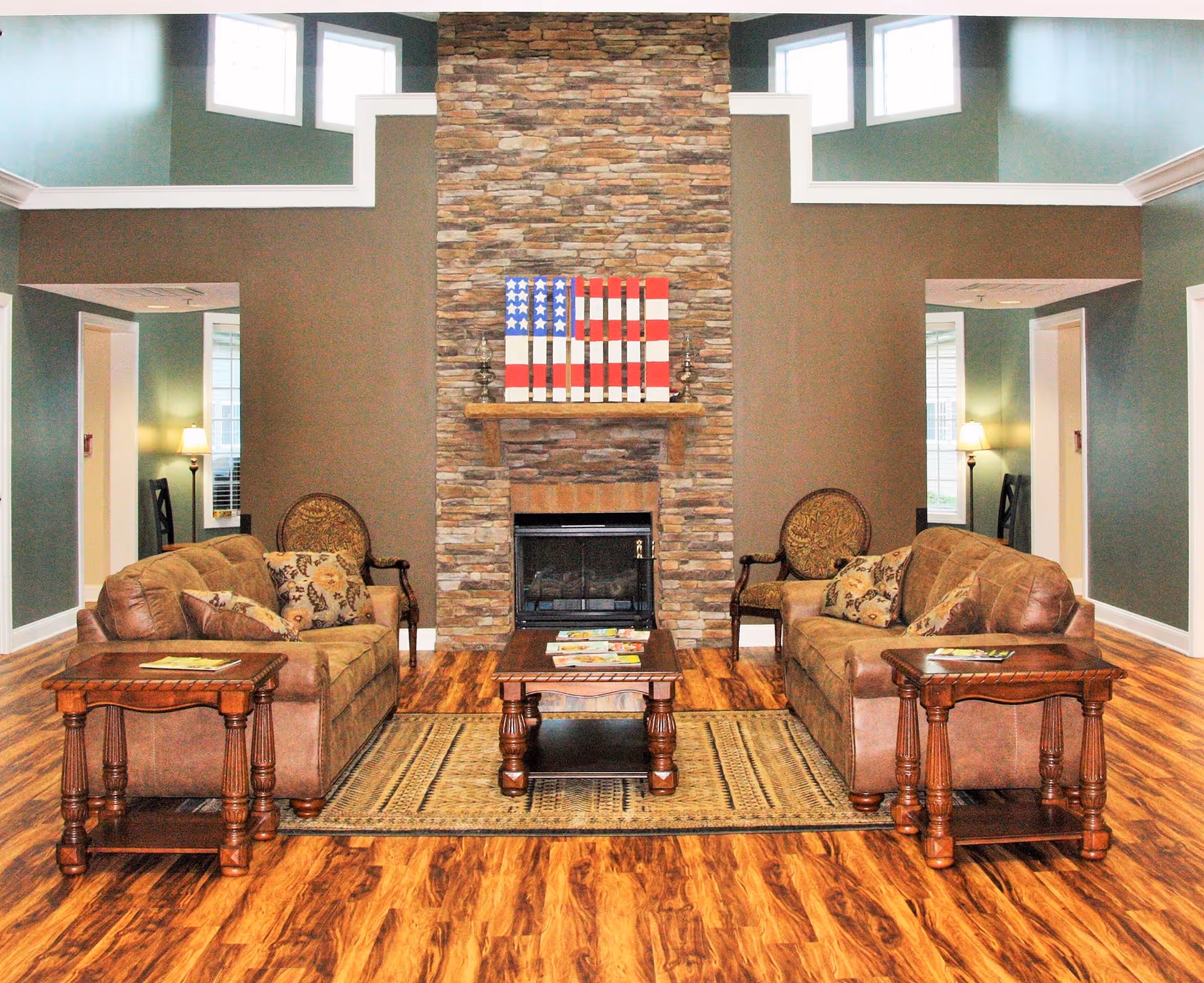 A cozy living room with two brown leather sofas facing each other, a wooden coffee table in the center on a patterned rug, and a stone fireplace with a wooden mantel decorated with an American flag artwork. The room has wooden flooring, green and brown walls, and windows near the ceiling letting in natural light.