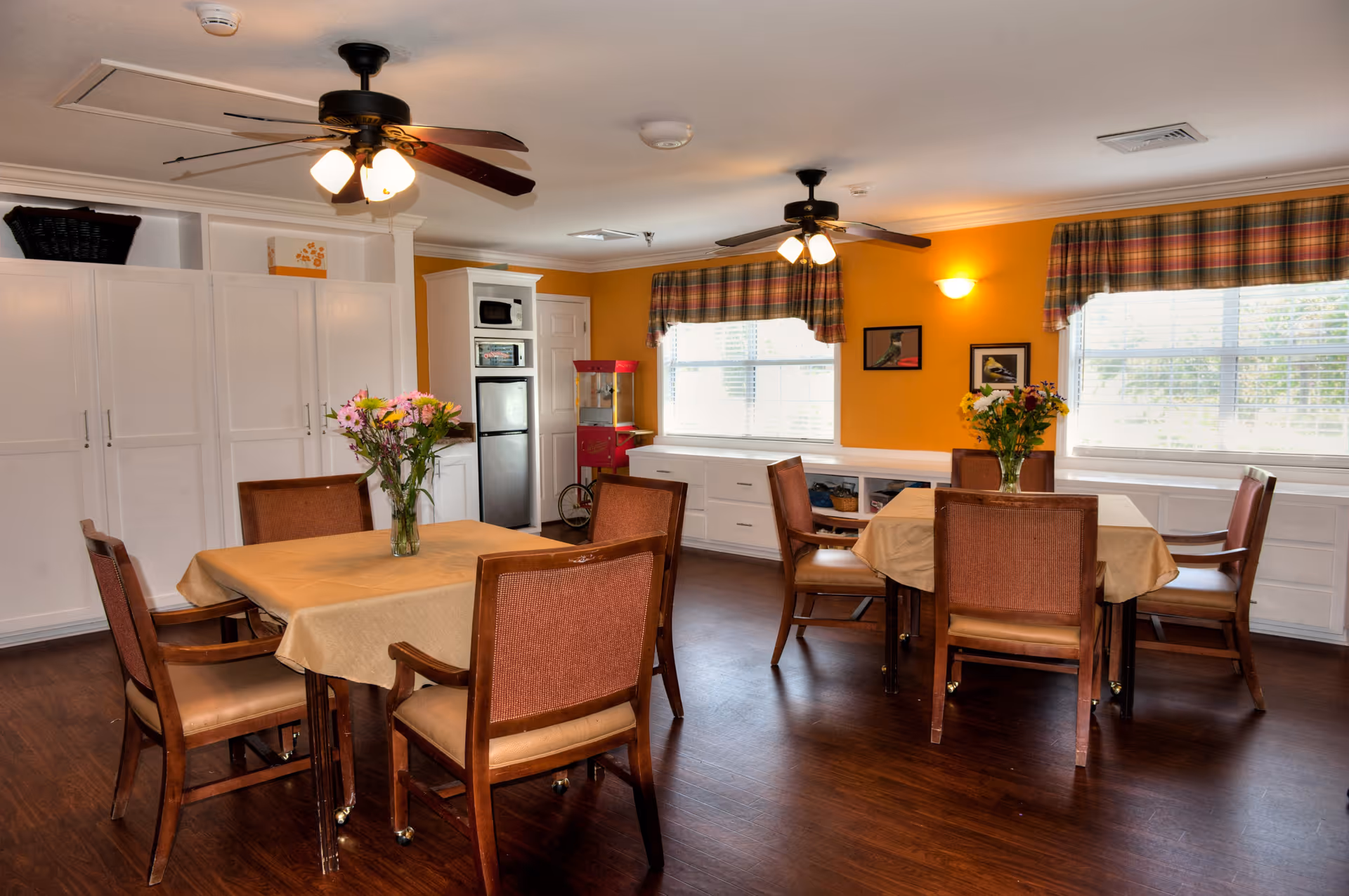 A cozy dining room with two square tables covered with beige tablecloths, each surrounded by four wooden chairs with brown cushions. Each table has a vase with fresh flowers. The room has warm yellow walls, two ceiling fans with lights, large windows with plaid valances, and white cabinetry along the walls. A small popcorn machine is visible near a white door.