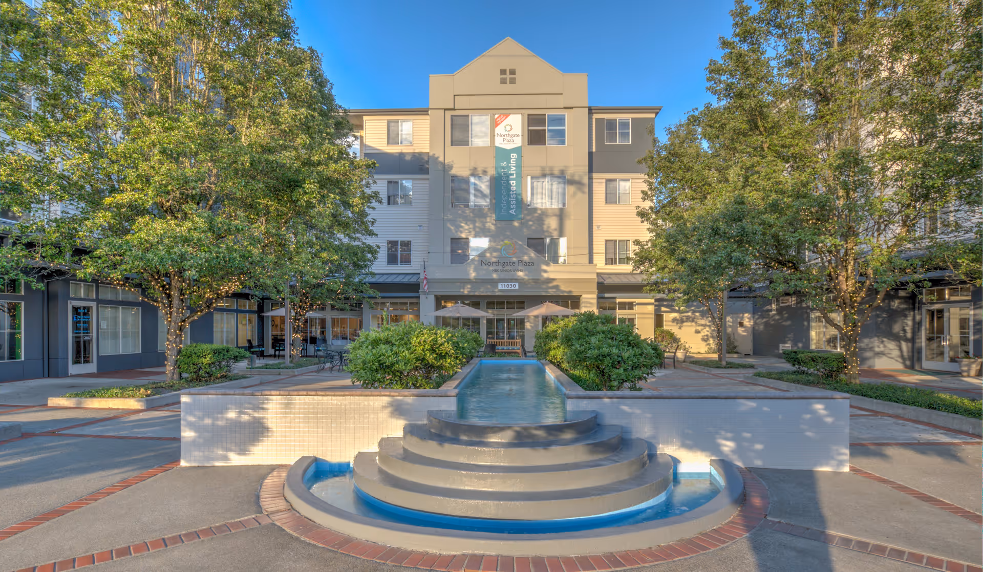 Front exterior view of Northgate Plaza, a multi-story assisted living facility, with a tiered circular water fountain in the foreground, surrounded by trees and bushes with string lights, and a paved courtyard area.