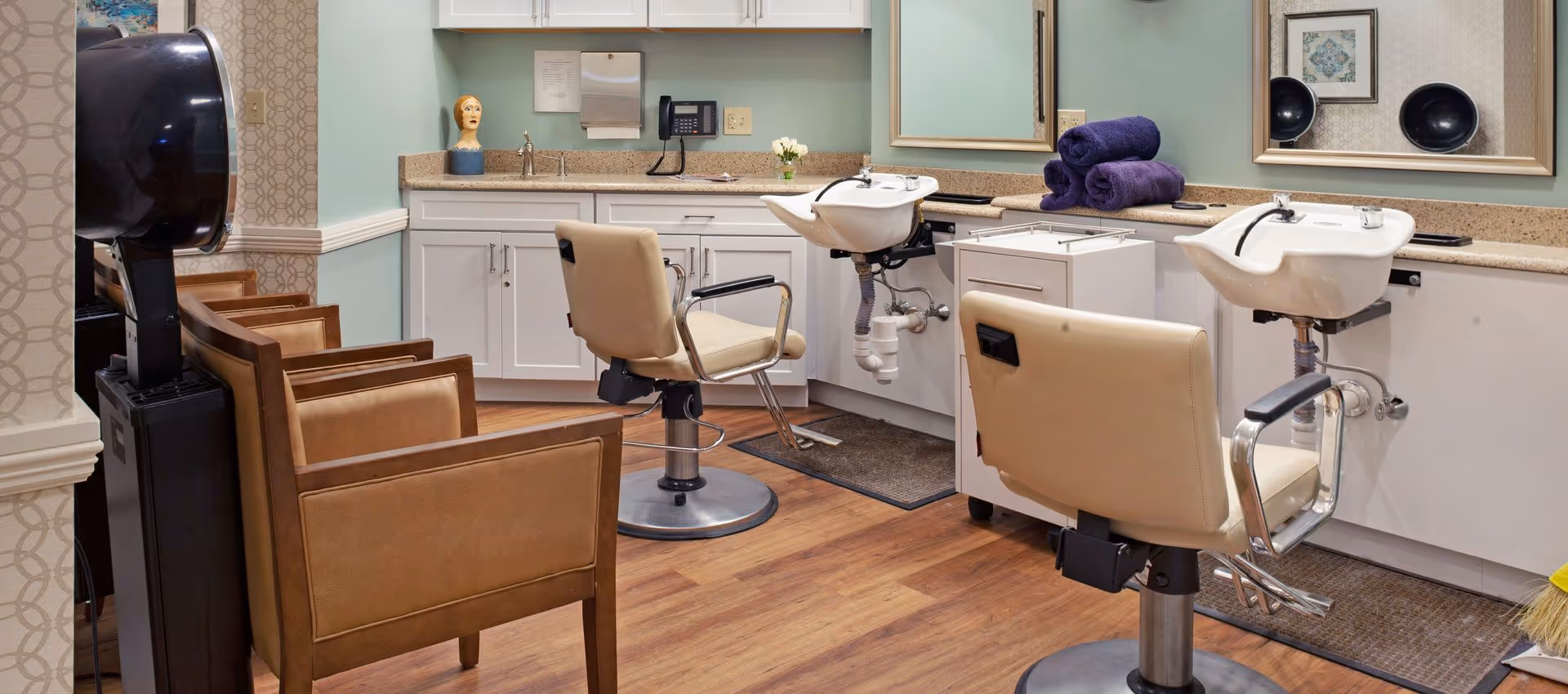 Interior view of a hair salon area in a senior living facility with two beige salon chairs in front of white sinks for hair washing, a counter with purple towels, a mirror, and additional seating with brown chairs. The room has light green walls and wood flooring.