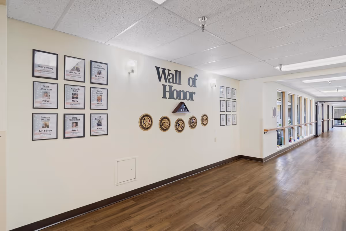 A hallway in Linda Vista Nursing and Rehab Center featuring a Wall of Honor display with framed photos and plaques honoring military veterans. The wall has the words 'Wall of Honor' prominently displayed along with military insignias and a folded American flag in a triangular case. The hallway has wood flooring, handrails along the windows, and bright overhead lighting.