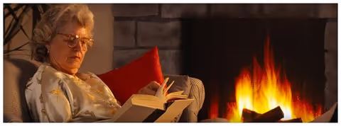 An older woman reading a book beside a glowing fireplace in a cozy living room.