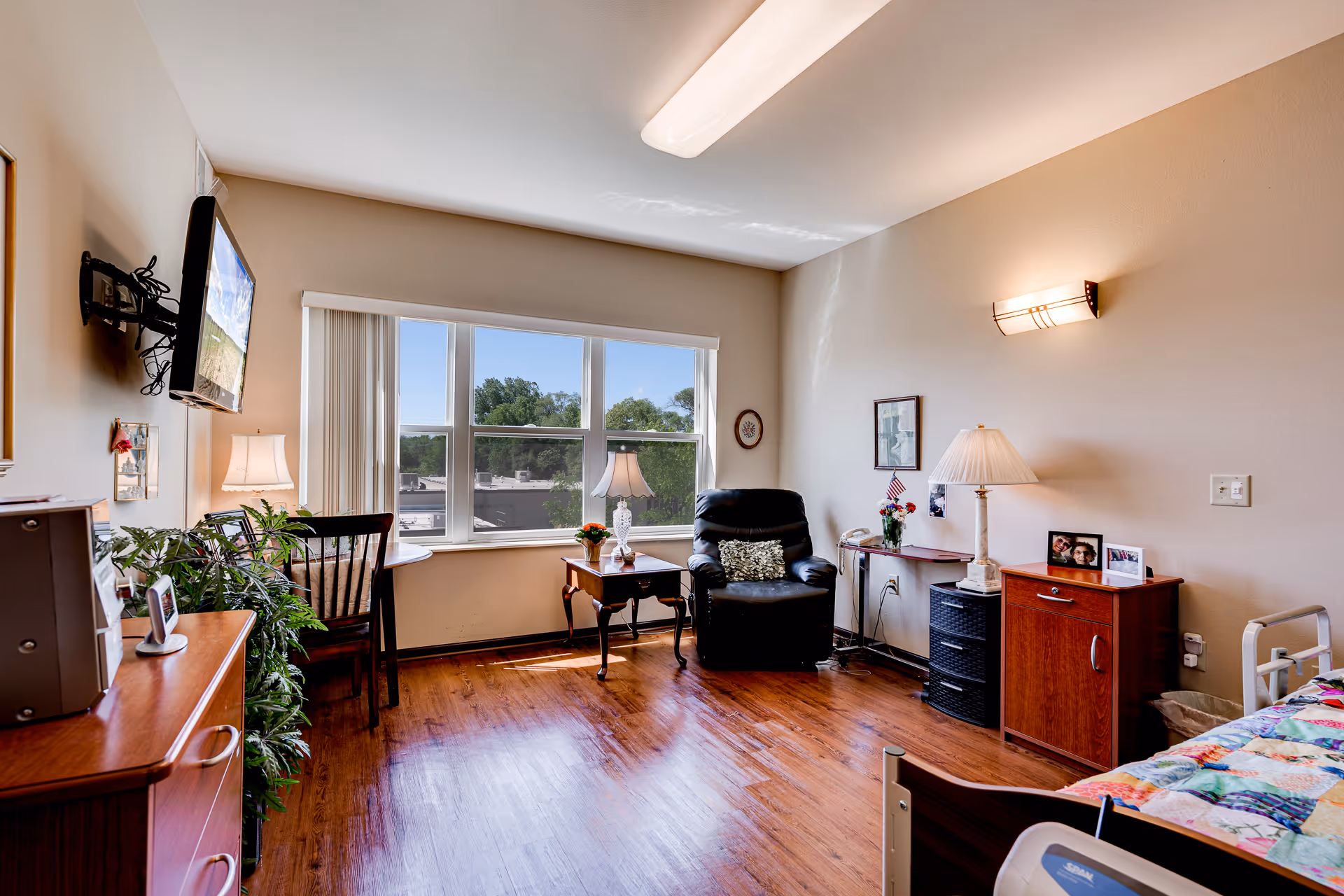 A bright and cozy senior living room with a large window letting in natural light. The room features a black recliner chair, a wooden side table with a lamp and flowers, a small desk with a chair, a wall-mounted TV, and a bed partially visible on the right side. The floor is wooden, and the walls are painted beige with framed pictures and a wall sconce light.