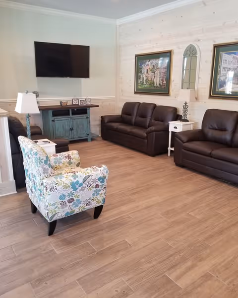 A cozy living room with two dark brown leather sofas and a floral patterned armchair arranged around a wooden floor. A flat-screen TV is mounted on the wall above a rustic teal cabinet. The walls are light-colored with wood paneling on one side, decorated with framed artwork and a decorative mirror. Two white side tables with lamps flank the sofas.