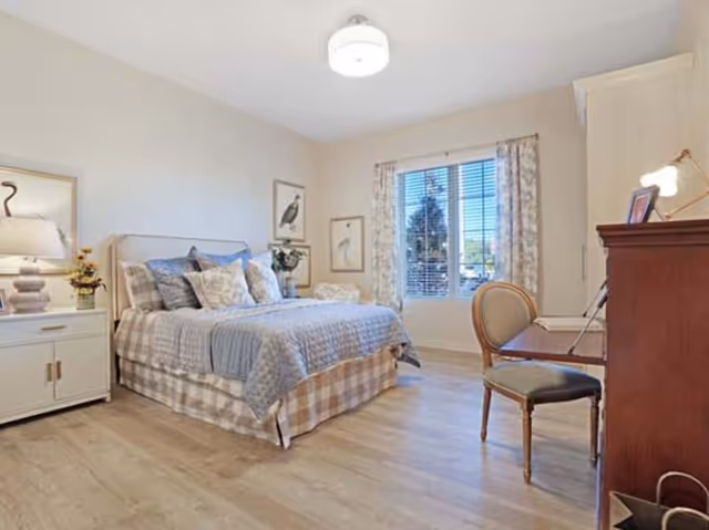 A bright and cozy bedroom with a large bed covered in patterned bedding and multiple pillows. There is a white nightstand with a lamp and flowers on the left side of the bed. The room has light-colored walls and wood flooring. A window with floral curtains lets in natural light. On the right side, there is a wooden desk with a chair and a small lamp.