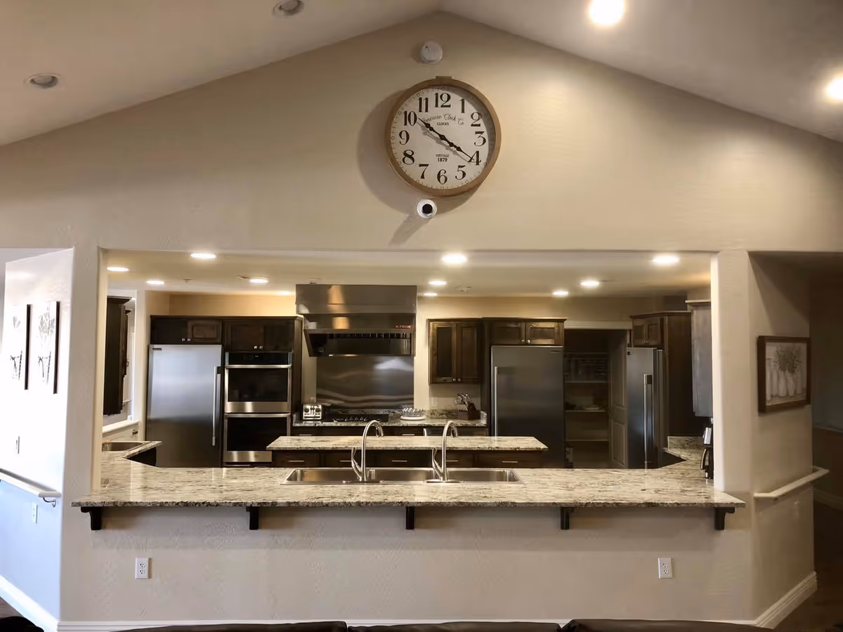 Modern kitchen area with granite countertops, stainless steel appliances including a refrigerator, oven, and stove. There is a double sink in the foreground and a large wall clock above the kitchen opening.