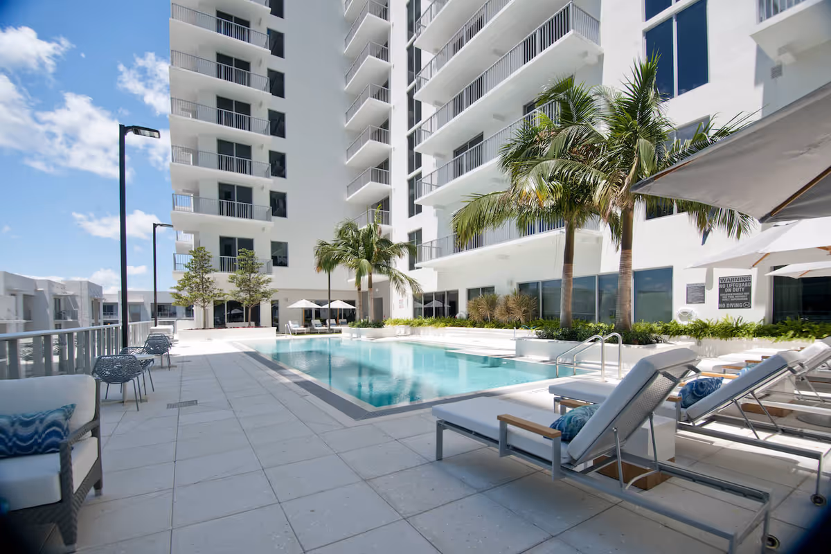 Outdoor swimming pool area at a senior living facility named Mirabelle, featuring lounge chairs with cushions, palm trees, umbrellas, and a modern white multi-story building in the background under a partly cloudy blue sky.