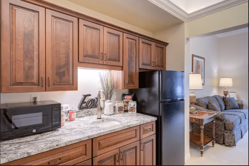 A kitchen area with wooden cabinets, a granite countertop, a black microwave, a black refrigerator, and a sink. Decorative items including a vase with tall grass and a sign that says 'Eat' are on the counter. In the background, a living room is visible with a patterned sofa, a wooden side table, two table lamps, and framed artwork on the wall.