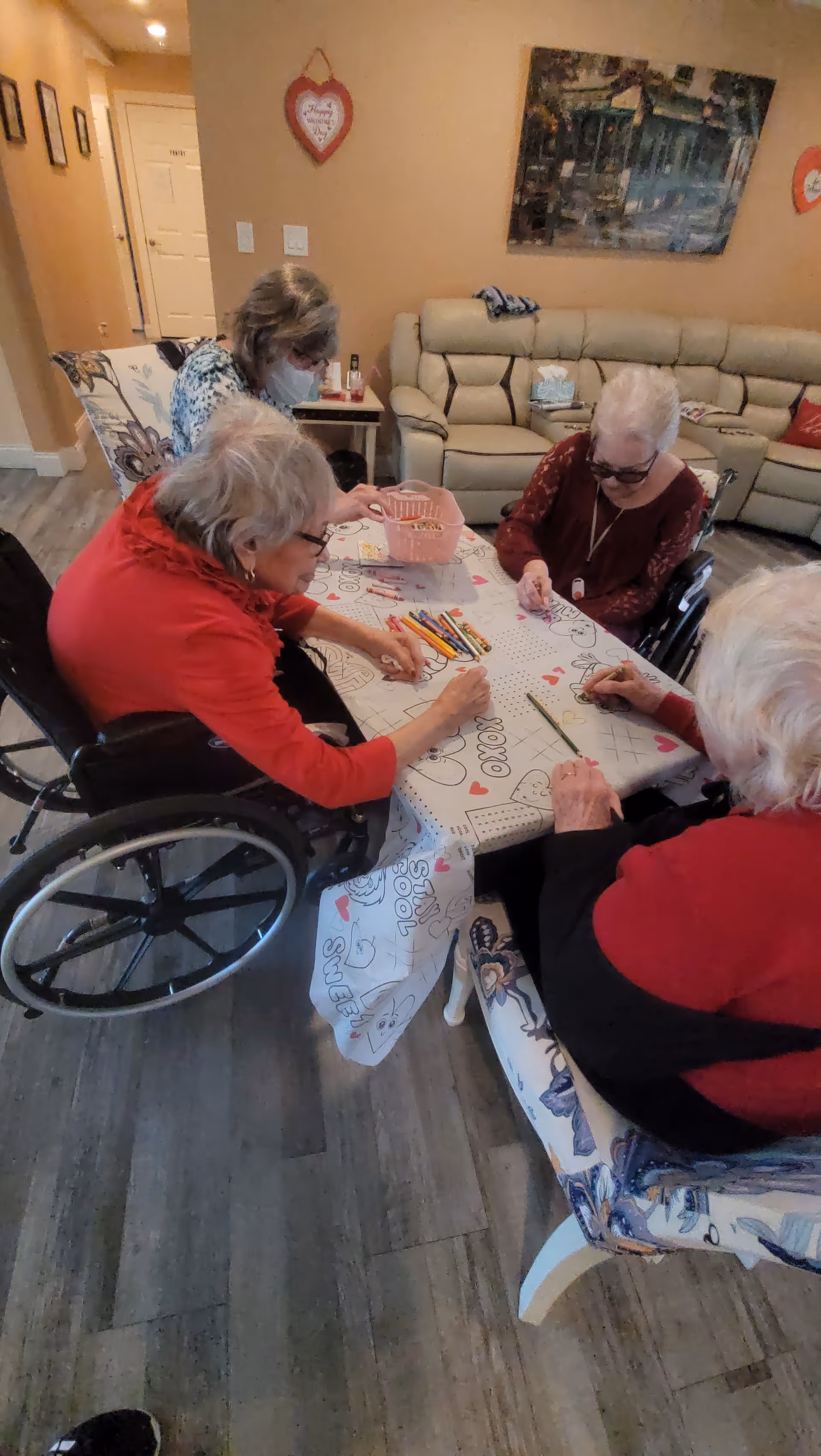 Four elderly women sitting around a table engaged in coloring activities with colored pencils. Two women are in wheelchairs, and the setting appears to be a cozy living room with a beige sectional sofa, wall decorations, and a wooden floor.
