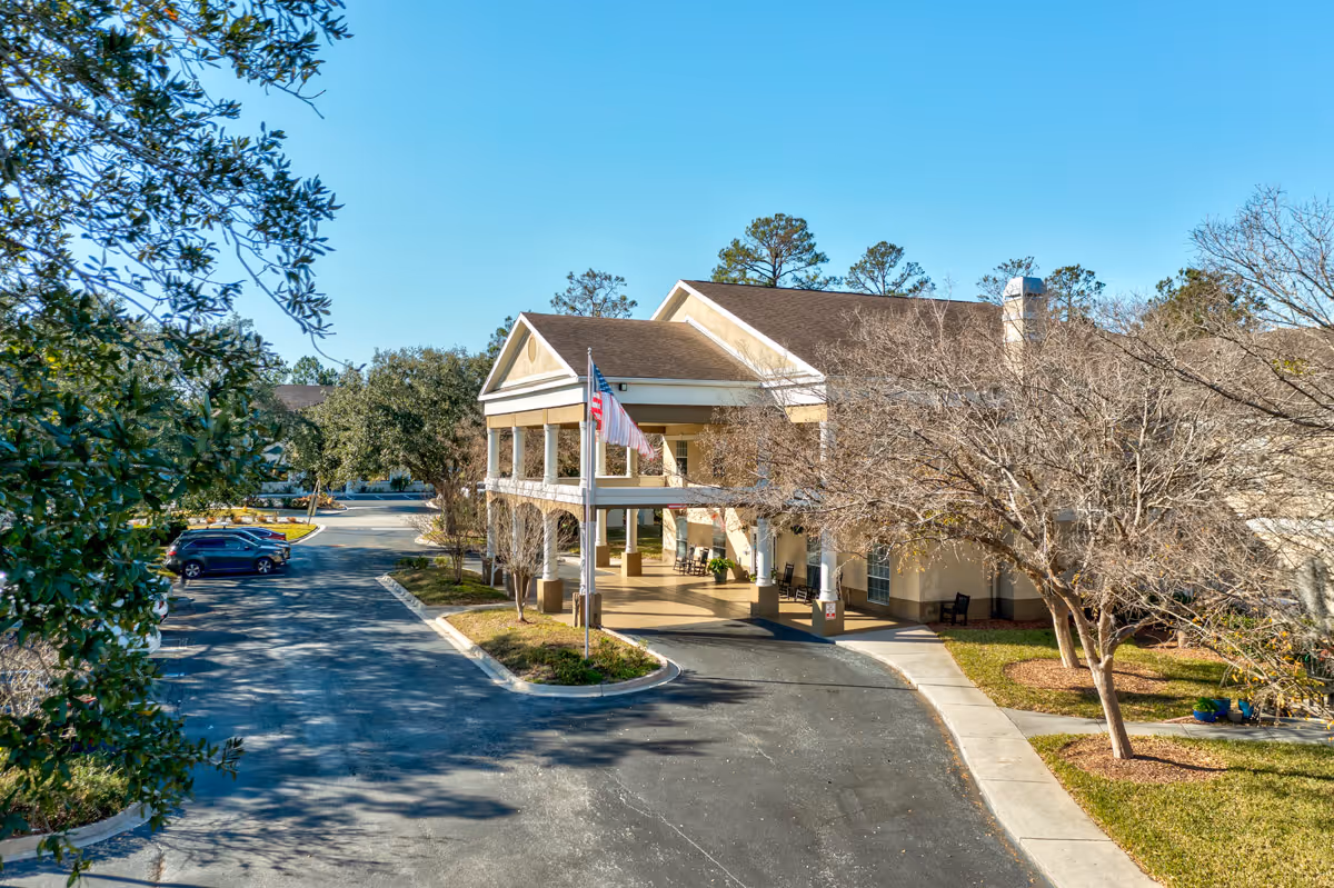 Exterior view of a senior living facility building with a covered entrance, an American flag, several trees including some without leaves, a parking lot with a few cars, and a clear blue sky.