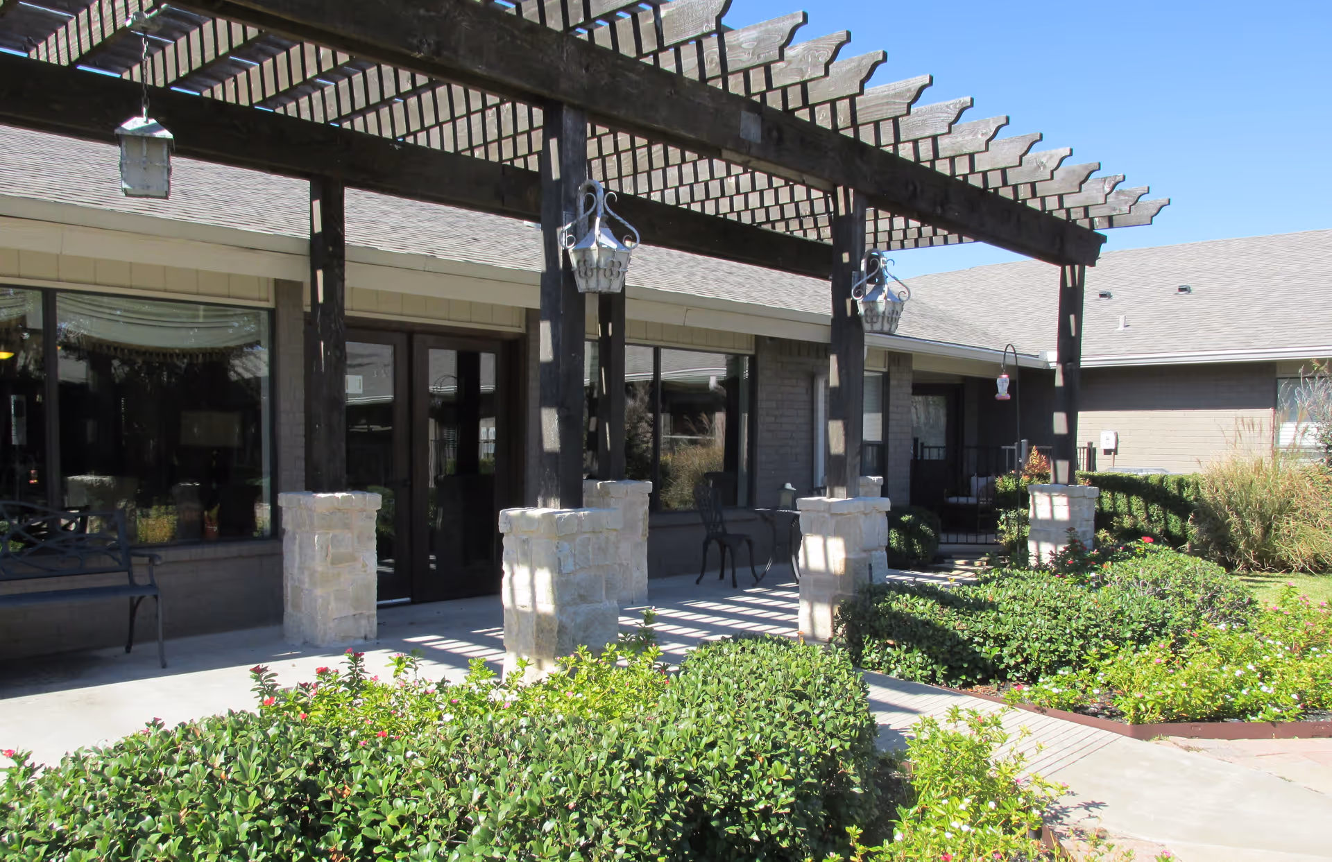 Outdoor patio area of a senior living facility with a wooden pergola supported by stone pillars, benches, chairs, and well-maintained green shrubs and plants under a clear blue sky.