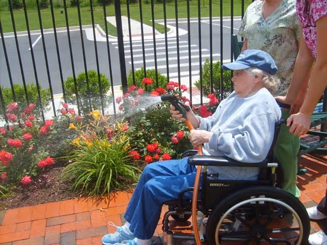 An elderly woman in a wheelchair waters a flower bed with red and yellow flowers using a garden hose, while two people stand behind her on a brick-paved area near a black metal fence and a road with a crosswalk.
