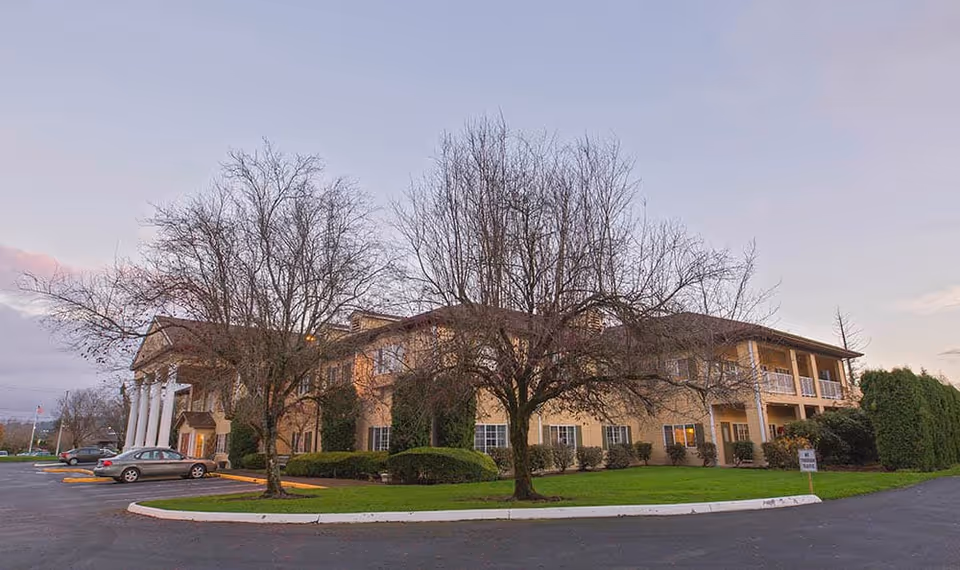 Front exterior of a two-story assisted living building with columns, leafless trees, parked cars, and a grassy lawn at dusk.