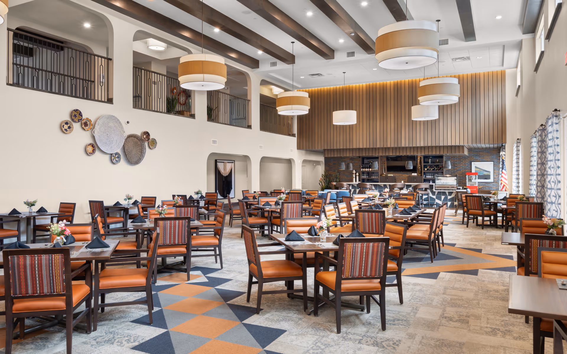 A spacious and well-lit dining room with multiple wooden tables and chairs arranged neatly. The chairs have orange cushions and patterned backs. The room features high ceilings with exposed wooden beams and large circular pendant lights hanging down. There is a decorative wall with woven baskets on one side and a serving area or bar with shelves and equipment at the far end. Large windows with patterned curtains allow natural light to fill the space.