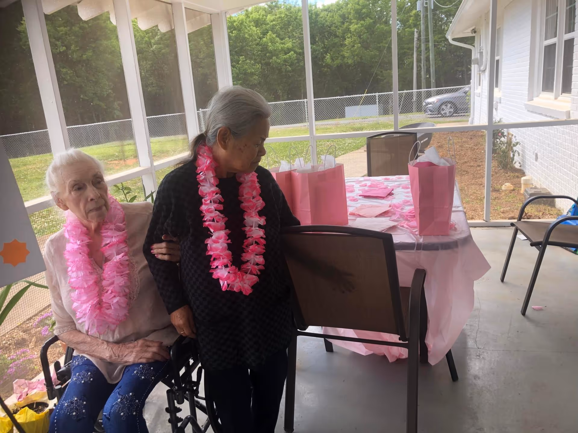 Two elderly women wearing pink flower leis are sitting and standing next to each other on a screened-in porch. One woman is seated in a wheelchair while the other stands beside her holding her arm. Behind them is a table covered with a pink tablecloth, pink gift bags, and pink decorations. The porch overlooks a fenced yard with trees and a parked car visible in the background.