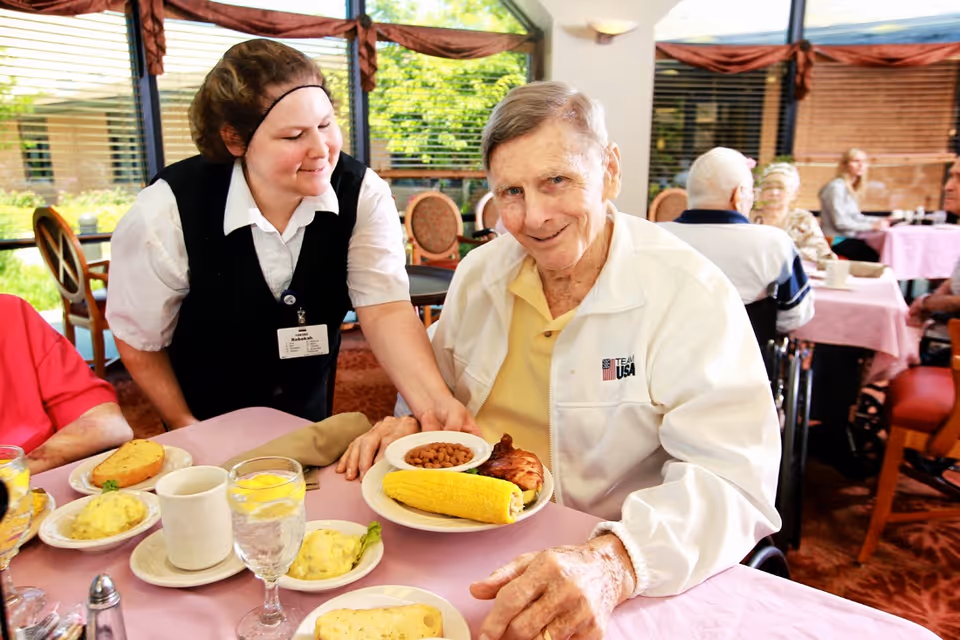 An elderly man in a white jacket sits at a dining table with a plate of food including corn on the cob, baked beans, and chicken. A smiling female staff member wearing a black vest and white shirt is serving him. Other elderly people are seated at tables in the background near large windows with greenery outside.