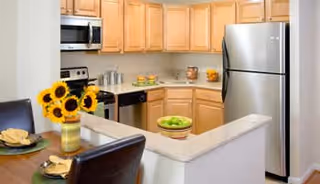 Modern kitchen with light wood cabinets, stainless steel refrigerator, microwave, and stove. A small counter area with a bowl of green apples and a vase of sunflowers is visible, along with two place settings on a breakfast bar with dark chairs.