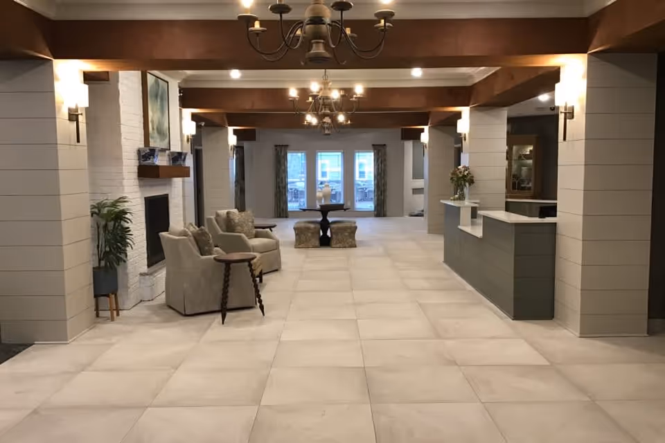 A spacious, well-lit common area with beige tiled flooring, light-colored walls with wooden beams on the ceiling, and multiple chandeliers. There are two armchairs with a small round wooden table between them near a white brick fireplace on the left. In the background, there is a round table with two cushioned stools near large windows with curtains. On the right side, there is a reception or service counter with a vase of flowers on it.
